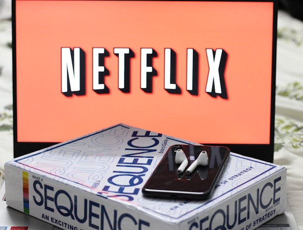 A screen displaying the Netflix logo in bold, black and white lettering against a vibrant red background. In front of the screen, there is a closed box for the board game 'Sequence', with a smartphone and wireless earbuds resting on top.