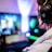 A person wearing headphones is sitting in front of a computer setup with multiple monitors displaying colorful, blurred graphics. The headphones have a distinctive logo illuminated in green. The ambient lighting and screen glow add to the immersive gaming environment.