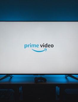 A television screen displays the Prime Video logo, situated on an entertainment unit in a dimly lit room. The room is illuminated with a subtle blue backlight, creating a calming atmosphere. Two small decorative items, resembling mushrooms, are placed on either side of the unit.