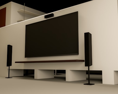 A modern home entertainment setup featuring a large flat-screen TV mounted on a light-colored wall. Two tall black speakers stand on either side of the TV. The room has a minimalist design with a clean, uncluttered look. Shelving and a wooden door are visible in the background.